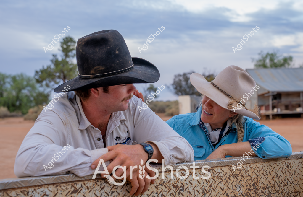 A man and a woman in classic rural Australian attire lean against a metal fence, sharing a moment of connection at what appears to be a remote cattle station. The man wears a black cowboy hat and a crisp white long-sleeved work shirt, while the woman sports a light-colored wide-brimmed hat and a bright blue button-up shirt. Their relaxed posture and proximity suggest a comfortable working relationship, typical of rural Australian agricultural settings.

The backdrop reveals a dry, sandy landscape with sparse vegetation and a tin-roofed building in the distance, characteristic of the Australian outback. Soft blue-gray clouds diffuse the light, creating a muted, expansive atmosphere that speaks to the vastness of rural agricultural landscapes. Their matching cowboy hats and lean against the textured metal fence highlight the shared culture and resilience of people working in remote agricultural environments.

The scene captures the essence of contemporary rural Australian life - connection, hard work, and an intimate understanding of the challenging but beautiful landscape they call home. Their stance suggests a moment of rest or conversation, perhaps discussing the day's work or upcoming tasks on the station.