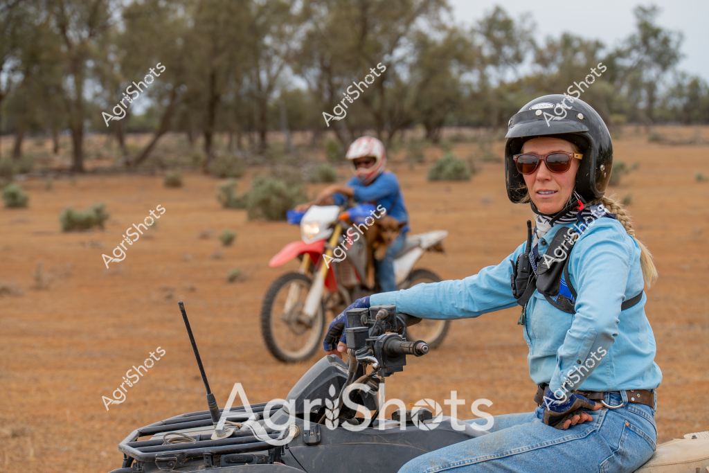 A woman rider sits confidently on an all-terrain vehicle (ATV) in a dry, sparse Australian outback landscape, wearing protective gear including a black helmet, sunglasses, and a light blue long-sleeved top. In the background, another rider on a red dirt bike navigates the arid terrain, which features scattered low shrubs and sparse eucalyptus trees across a reddish-brown soil surface. The scene captures the rugged nature of rural Australian terrain, with both riders equipped for off-road exploration, highlighting the robust spirit of outdoor adventure in a challenging landscape. The woman's positioning on the ATV suggests she is experienced and comfortable in this remote environment, with her sunglasses and helmet providing protection from the harsh Australian sun and dust. The sparse vegetation and open ground underscore the challenging conditions of this remote agricultural or pastoral setting.