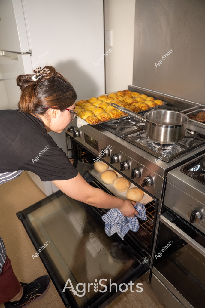 A woman stands at a gas stove preparing freshly baked pastries, carefully removing golden-brown rolls from the oven using a checkered kitchen towel. The kitchen is modern with a stainless steel stove, and multiple trays of yellow-golden pastries are visible on the stovetop. She wears a black t-shirt and has her hair pulled up in a bun, concentrating intently on removing the hot baked goods. A silver pot sits on one of the stove burners, suggesting she is in the midst of a baking or cooking process. The scene captures a moment of culinary preparation, with warm, appetizing colors and the careful movements of someone skilled in baking.