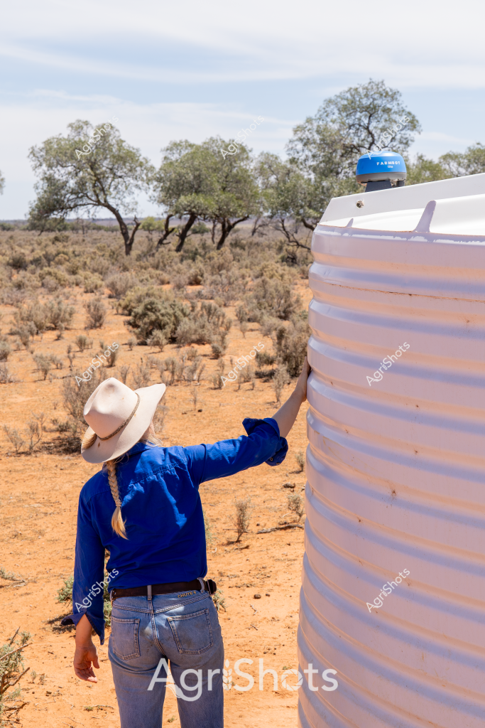 A woman wearing a wide-brimmed sun hat and bright blue long-sleeved shirt stands beside a large white water storage tank in an arid Australian landscape. The background reveals sparse, weathered trees and low scrubby bushes characteristic of an inland pastoral region, with reddish-brown soil and muted grey-green vegetation stretching to the horizon. She appears to be checking or maintaining the water tank, which has a blue sensor or monitoring device mounted on top, critical infrastructure for managing water resources in a dry environment. The scene captures the resilience and practical necessities of rural Australian agricultural life, where water management is essential for survival. The stark landscape and solitary figure highlight the challenging conditions faced by farmers and agricultural workers in remote areas, where each water resource is precious and carefully monitored.