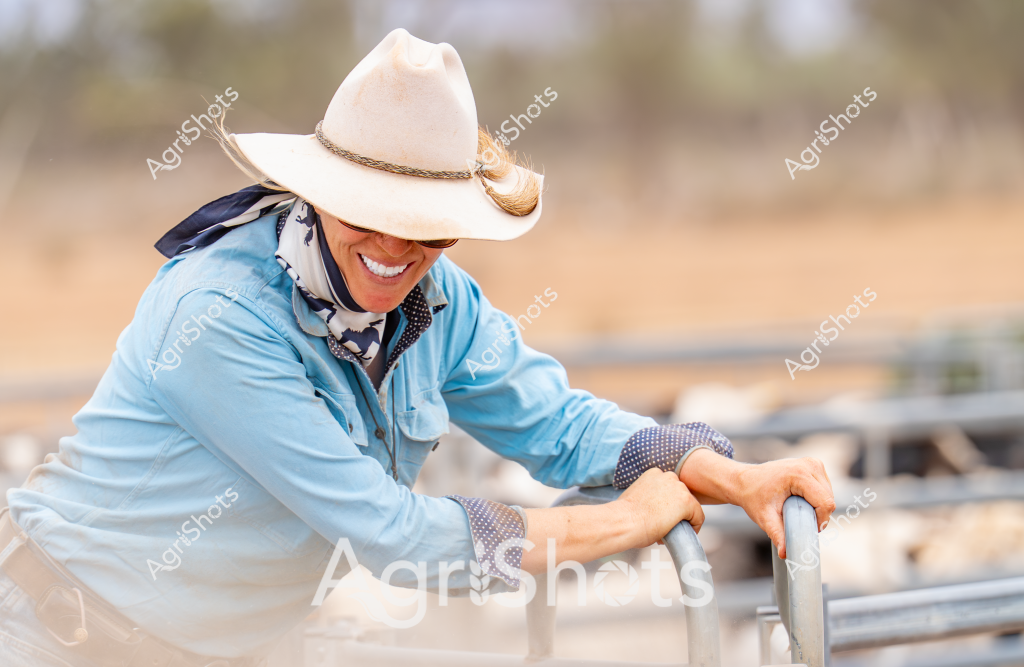 A woman farmer in a light blue shirt and white cowboy hat leans confidently over a metal livestock fence, her warm smile and relaxed posture capturing the spirit of rural life. She wears polka-dotted work gloves and a patterned neckerchief, suggesting preparedness for farm tasks. The background reveals a soft, blurred landscape of dusty paddocks and scattered trees, typical of an Australian rural setting. Her cowboy hat is angled to shade her eyes, and her leaning posture suggests an easy familiarity with the farm environment. The image radiates a sense of joy and connection to the land, embodying the resilience and optimism of Australian agricultural workers.