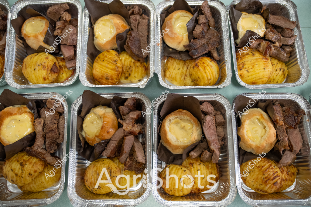 Multiple aluminium takeaway containers display identical gourmet meal preparations featuring roasted meat cuts, golden hasselback potatoes, and golden pastry items. The carefully portioned meals showcase precise culinary presentation with each container containing identical protein, potato, and pastry components arranged systematically. The potatoes feature distinctive accordion-style slicing with golden-brown surfaces, while the meat appears to be cubed or sliced beef portions with a rich brown caramelized exterior. A golden pastry item - potentially a Yorkshire pudding or cheese-topped bread roll - sits prominently in each container, complementing the protein and potato elements. The uniformity suggests these are pre-prepared meal packages, likely for catering, meal delivery, or restaurant takeaway services, demonstrating professional kitchen precision and portion control.