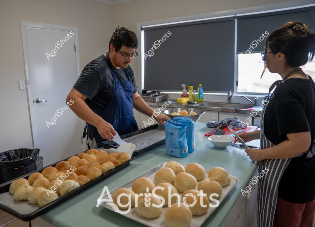 A man and a woman are working together in a kitchen, preparing a batch of freshly baked bread rolls. The man, wearing a blue apron and glasses, is carefully handling golden-brown bread rolls on large baking trays, while the woman in a black t-shirt stands nearby, observing the process. Multiple trays of perfectly shaped, uniformly colored bread rolls are laid out on the kitchen counter, suggesting a professional or commercial baking operation.

The kitchen environment is clean and modern, with neutral-toned walls, a window with roller blinds, and a stainless steel countertop. Cooking ingredients and supplies are visible, including a blue bag of what appears to be flour and some bottles in the background. The precise arrangement of the bread rolls and the careful positioning of the bakers indicate a methodical approach to bread production.

The scene captures the collaborative nature of food preparation, with both individuals focused on their tasks. The warm, golden color of the bread rolls creates a sense of comfort and appetite appeal, highlighting the craft of artisan bread making. The image could represent a small bakery, commercial kitchen, or professional cooking environment dedicated to producing high-quality baked goods.