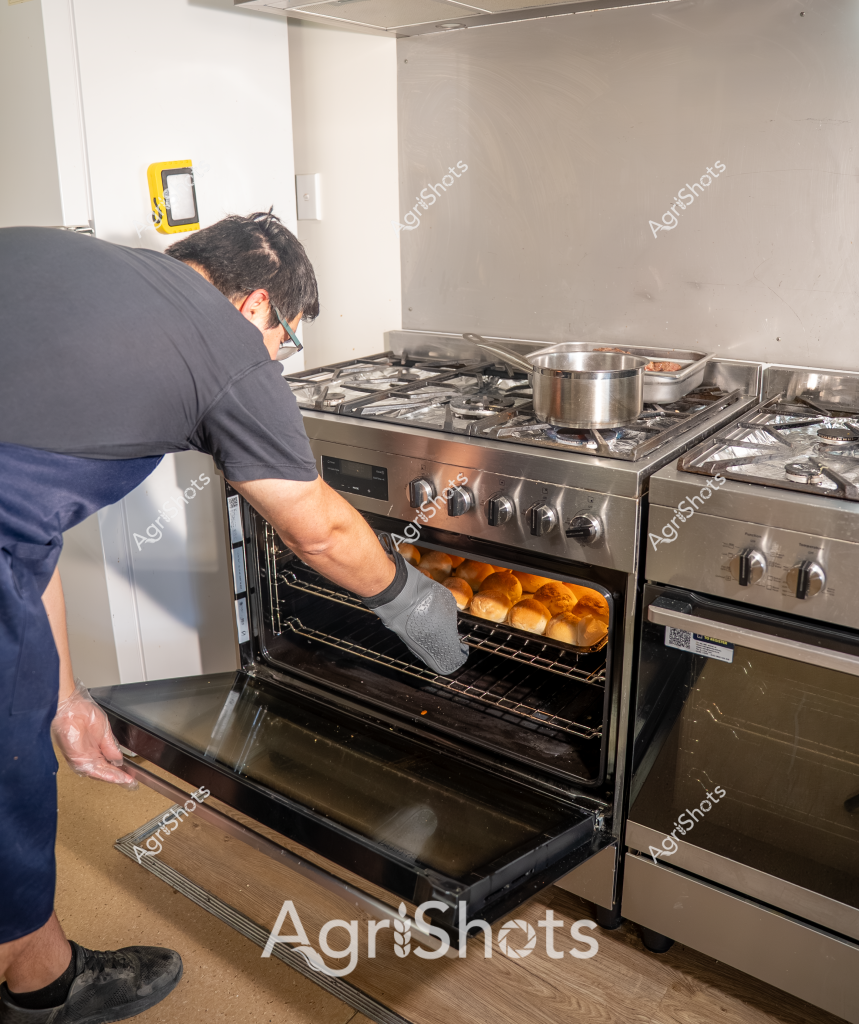 A professional chef in a grey shirt and navy apron carefully removes freshly baked golden-brown bread rolls from a stainless steel oven in a commercial kitchen. The oven is part of a professional-grade gas stove with multiple burners, and a stainless steel pot is visible on the stovetop. The baker is wearing a protective grey oven mitt and leaning carefully to extract the perfectly browned bread, demonstrating precision and skill in food preparation. The kitchen environment appears clean and organized, with white walls and a professional-grade cooking setup that suggests a high-end culinary setting. The warm, golden color of the bread rolls contrasts beautifully with the metallic surfaces of the kitchen equipment, creating an appealing visual of fresh baking in progress.