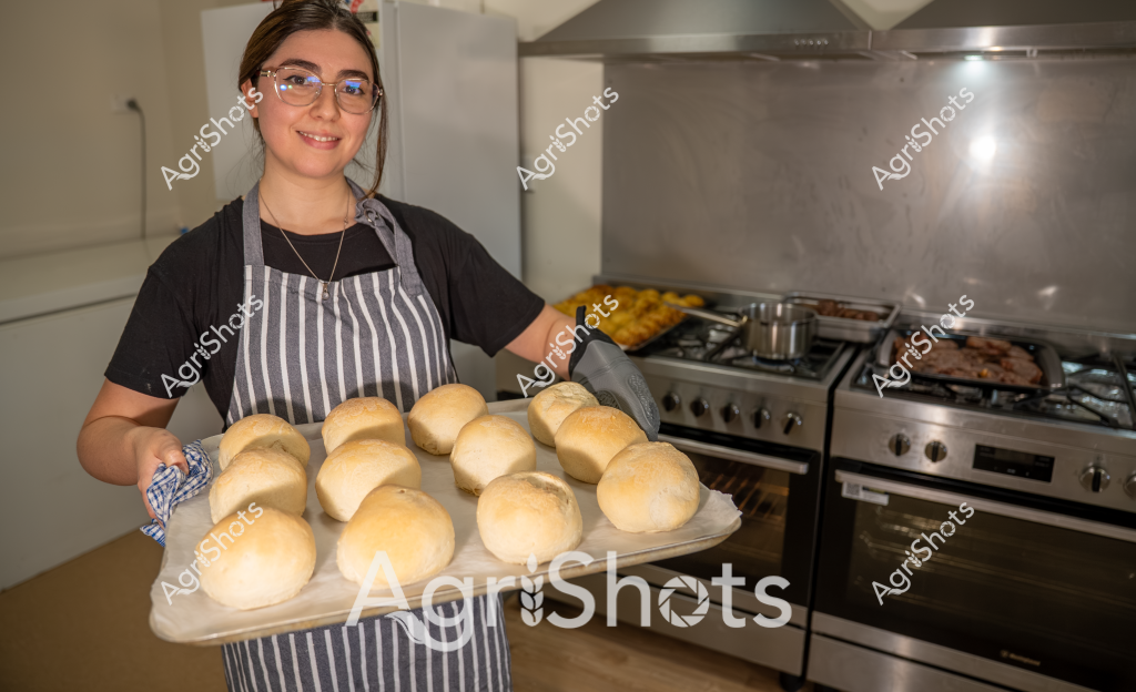A woman baker stands in a home kitchen, proudly displaying a freshly baked batch of golden bread rolls on a baking tray. Wearing a striped gray apron over a black t-shirt and blue-rimmed glasses, she smiles warmly while holding the tray of perfectly round, uniformly risen bread rolls. Behind her, a professional stainless steel stove and kitchen range provide the backdrop, with additional cooking items and trays visible. The bread rolls have a beautiful golden-brown crust, suggesting they've just come out of the oven and are at the peak of freshness. Her neat, pulled-back hair and confident pose indicate a skilled and practiced baker who takes pride in her craft of bread-making.