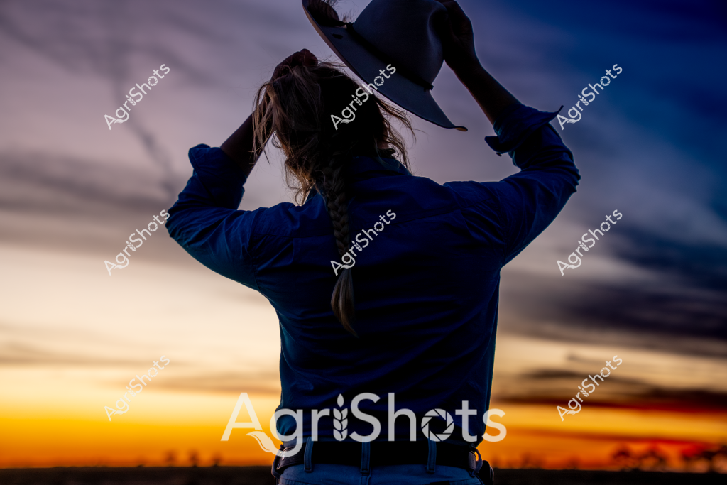 A woman stands silhouetted against a dramatic sunset sky, her back to the camera, wearing a deep blue jacket. She raises her hands above her head, creating a sense of freedom and contemplation as the horizon blazes with warm orange and golden hues transitioning to deep blue. The sky is filled with soft, sweeping clouds that create a sense of expansive rural atmosphere, suggesting an agricultural landscape at the day's end. Her ponytailed hair moves slightly, indicating a gentle breeze across what appears to be an open paddock or farm setting. The image captures a moment of quiet reflection, embodying the solitude and connection to landscape that characterizes rural Australian experience.