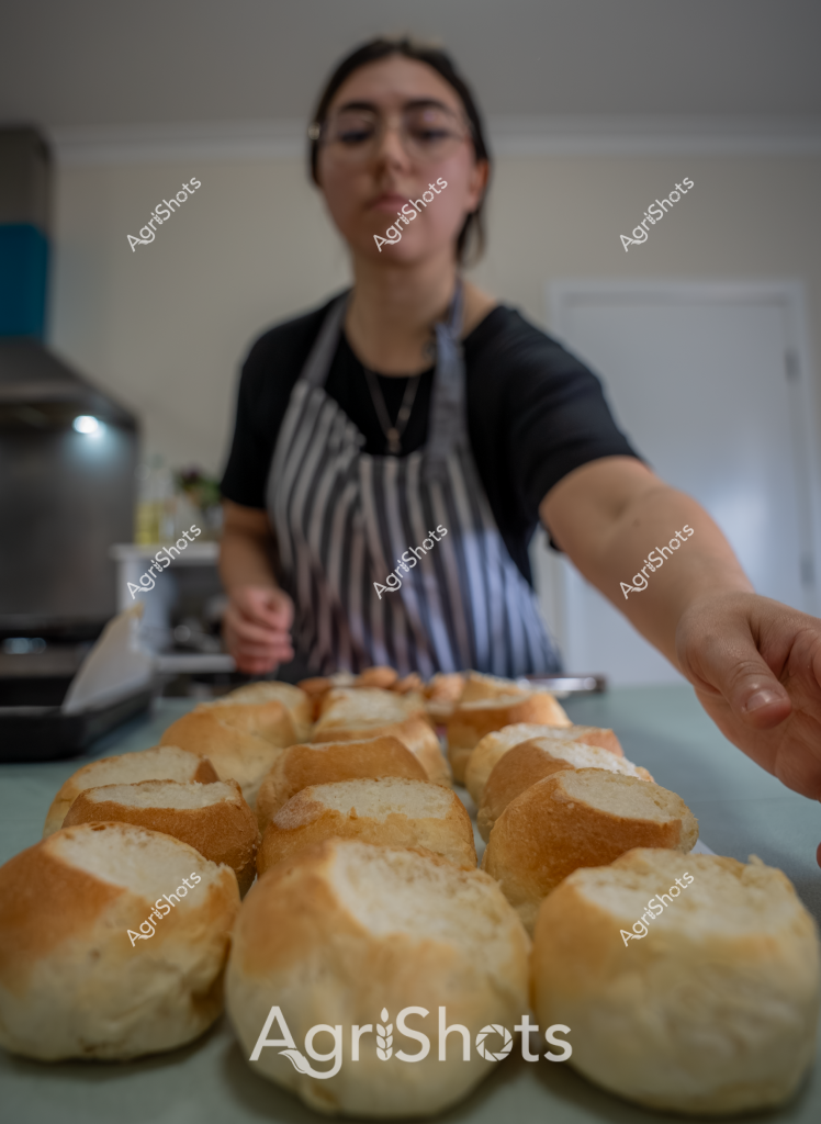 A woman in a striped apron stands in a kitchen, carefully arranging freshly baked bread rolls on a kitchen counter. Her eyes are closed, suggesting she is appreciating the aroma of the warm, golden-brown bread. The bread rolls have a uniform shape and a glossy, slightly crisp-looking crust, indicating they have just been removed from the oven. The kitchen environment is clean and bright, with soft lighting highlighting the texture of the bread. The scene captures a moment of culinary craftsmanship, showcasing the care and skill involved in home baking. The warm, inviting atmosphere evokes the comfort and satisfaction of freshly made bread.