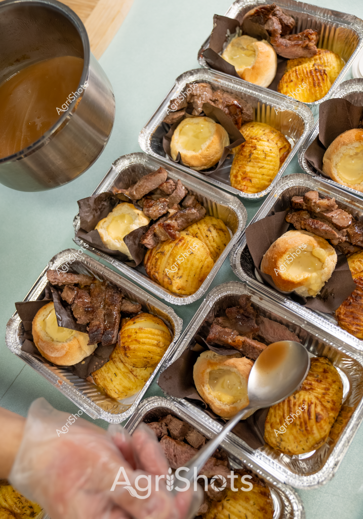 A professional catering setup showcases precisely portioned meat dishes prepared in individual aluminum takeaway containers. Each container features tender, browned meat portions alongside golden, crisp roasted potatoes and a freshly baked bread roll. A large stainless steel pot stands nearby, suggesting professional food preparation for an event or catering service. The meal portions are uniformly arranged, demonstrating meticulous culinary precision and portion control. The gleaming metallic surfaces and carefully plated components highlight the attention to detail in this commercial food service preparation.

The image captures a moment of professional food service organization, with multiple identical meal trays laid out systematically. The meat appears to be slow-cooked or braised, with a rich brown color indicating careful seasoning and cooking technique. The potatoes are sliced and roasted to a uniform golden-yellow, showing expert kitchen preparation. A gloved hand is visible in the frame, suggesting ongoing food service preparation or portioning.