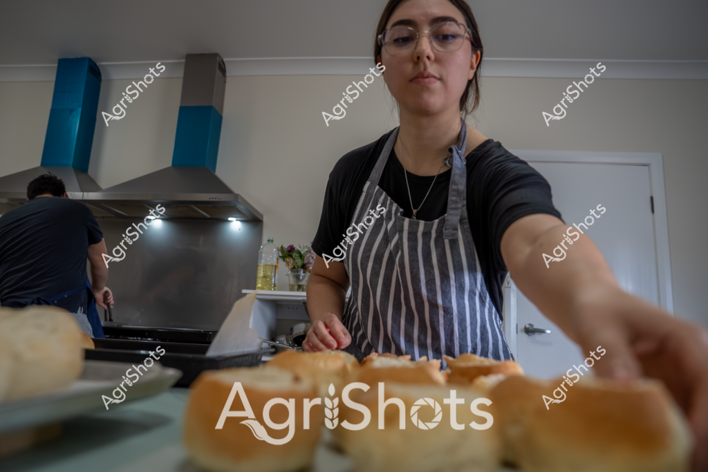 A woman wearing a striped apron and glasses prepares food in a contemporary kitchen, standing near a stainless steel range hood. In the foreground, several bread rolls or buns are visible, suggesting she is in the process of preparing a meal or sandwich. Another person is partially visible working in the background near the cooking area. The kitchen has a clean, modern aesthetic with white walls and sleek appliances. Her posture and focused expression indicate she is carefully preparing food, with precise and deliberate movements.

The scene captures an everyday culinary moment, highlighting the preparation of what appears to be fresh bread or bakery products. The neutral color palette of the kitchen - whites, grays, and blacks - creates a calm, professional cooking environment. Her gray and white striped apron suggests she is taking her food preparation seriously, whether for personal or professional purposes.