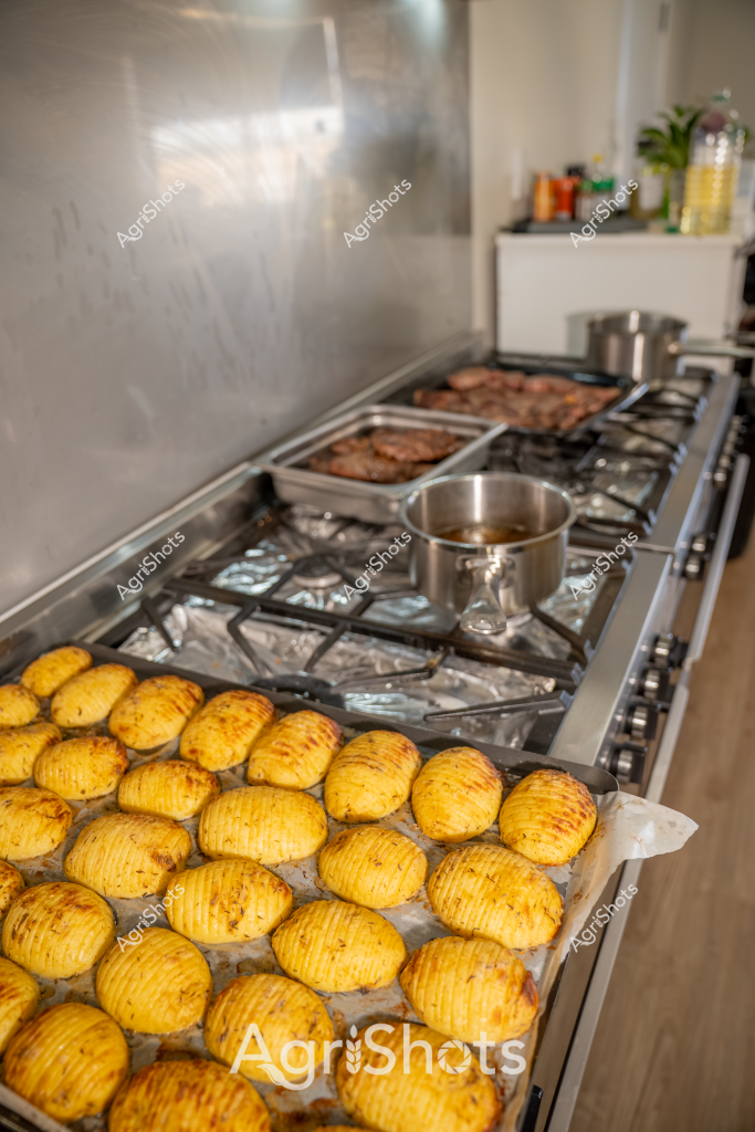 A bustling commercial kitchen scene captures a series of golden hasselback potatoes lined up meticulously on a stainless steel stovetop. The vibrant yellow-golden potatoes have been carefully sliced in an accordion style, creating an intricate texture that allows for crispy edges and even cooking. Nearby, some meat cuts are sizzling in stainless steel trays on the gas burners, suggesting a professional food preparation environment. A small stainless steel pot sits on the stovetop, likely for preparing sauce or additional ingredients. The kitchen backdrop reveals a clean, professional workspace with neutral tones and glimpses of cooking ingredients and a small plant, indicating a well-organized culinary setting. The carefully prepared potatoes showcase precision and attention to detail in food preparation, highlighting the artistry of professional cooking techniques.