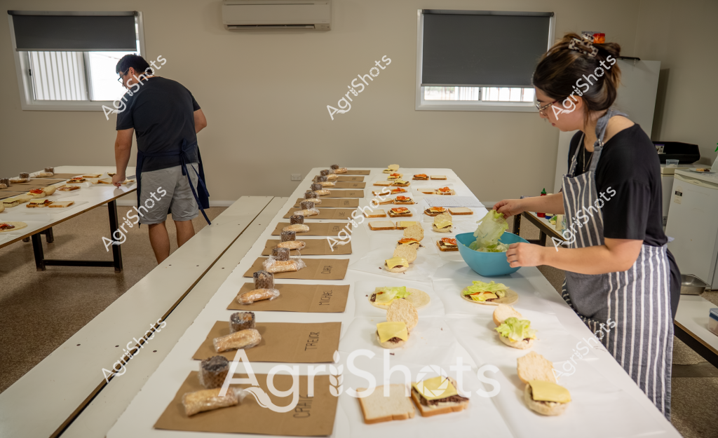 A woman in a striped apron and glasses prepares food items in what appears to be a professional food preparation or testing kitchen. Multiple sandwiches, bread slices, and ingredient containers are meticulously arranged across long white tables. Brown paper labels are placed systematically beside different food components, suggesting a carefully organized culinary experiment or sensory evaluation process.

A man in a dark shirt and gray shorts stands at the far end of the workspace, also engaged in food preparation. The room has a clean, clinical atmosphere with white walls, window blinds, and a mounted air conditioning unit, indicating a controlled environment for food research or product development.

The scene looks like a professional food testing setup, with each sandwich or ingredient precisely positioned. The methodical arrangement suggests a scientific approach to food preparation, possibly for taste testing, recipe development, or quality control in a commercial food production setting.

The neutral color palette - whites, grays, and browns - emphasizes the clinical, systematic nature of the workspace. The individuals are focused and deliberate in their actions, working with precision and attention to detail across the expansive preparation area.