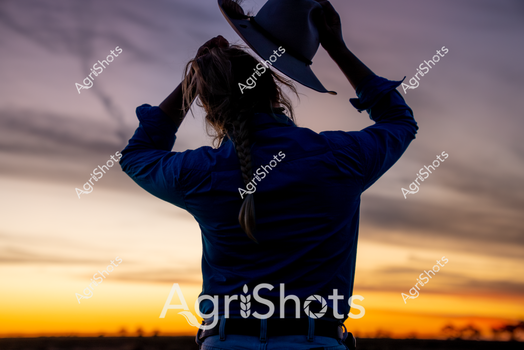 A woman wearing a blue jacket stands with her back to the camera, silhouetted against a dramatic sunset sky. Her hair is pulled back in a ponytail, and she is holding her hat above her head, creating a striking profile against the golden and purple horizon. The landscape stretches out behind her, with distant trees marking the edge of the sky and land. The lighting captures a moment of quiet reflection, with warm sunset colors creating a sense of peace and contemplation in the rural setting. The image evokes a sense of solitude and connection to the landscape, typical of rural Australian experience.