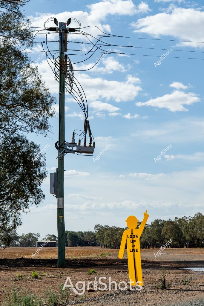 A striking safety awareness scene highlighting electrical hazard risks in rural Australia. A bright yellow silhouette of a safety figure stands near a power pole, with its "Look Up and Live" warning dramatically positioned against a backdrop of dry agricultural land and eucalyptus trees. The green power pole rises sharply against a blue sky with soft white clouds, its complex network of electrical cables and transformers demonstrating the intricate infrastructure that delivers electricity to remote farming regions. Sparse ground vegetation and a compacted dirt area surrounding the pole emphasize the harsh, arid landscape typical of Australian rural environments. The yellow safety figure's raised arm points upward, serving as a critical reminder of the potential dangers of electrical infrastructure and the importance of maintaining safe distances around power equipment.