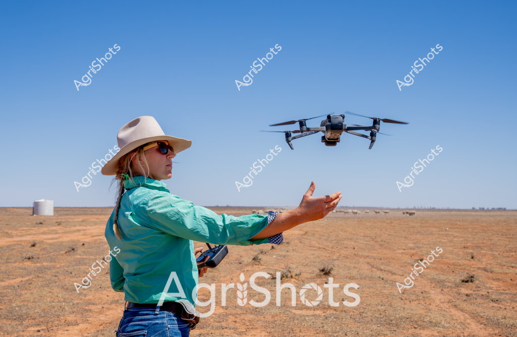 A woman in a turquoise long-sleeved shirt and wide-brimmed sun hat operates a drone in a stark, arid Australian landscape. Standing amid reddish-brown earth and sparse saltbush, she extends her hand to launch or guide a quadcopter drone, likely conducting agricultural surveying or monitoring. Wearing sunglasses to protect against the intense outback sunlight, she holds a remote control, demonstrating modern precision agricultural technology in a remote, dry environment. A white water tank is visible in the distant background, suggesting this is a rural station or farm property. The clear blue sky and open terrain highlight the expansive nature of Australian agricultural landscapes, where technological solutions are increasingly used to manage and understand challenging environmental conditions.