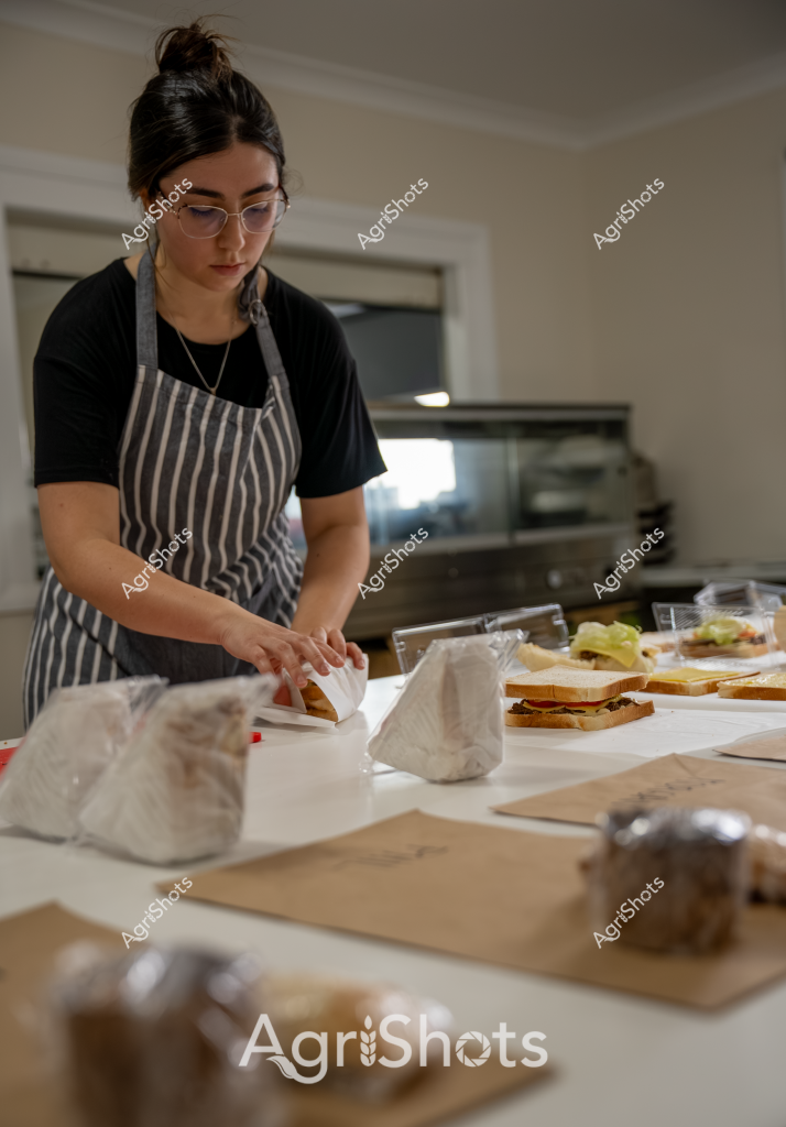 A woman wearing glasses and a gray and white striped apron carefully prepares sandwiches in a kitchen setting. She stands at a clean white countertop, surrounded by several wrapped sandwich packages and brown paper bags, suggesting a commercial food preparation environment. Her dark hair is pulled back in a neat bun, and she focuses intently on her work, meticulously arranging or packaging the sandwiches with precision. The kitchen appears professional and well-organized, with a stove visible in the background and soft, neutral lighting creating a calm atmosphere. Several completed sandwiches are laid out on the counter, wrapped in white paper, indicating a systematic approach to food production, potentially for a cafe, catering service, or meal delivery business.