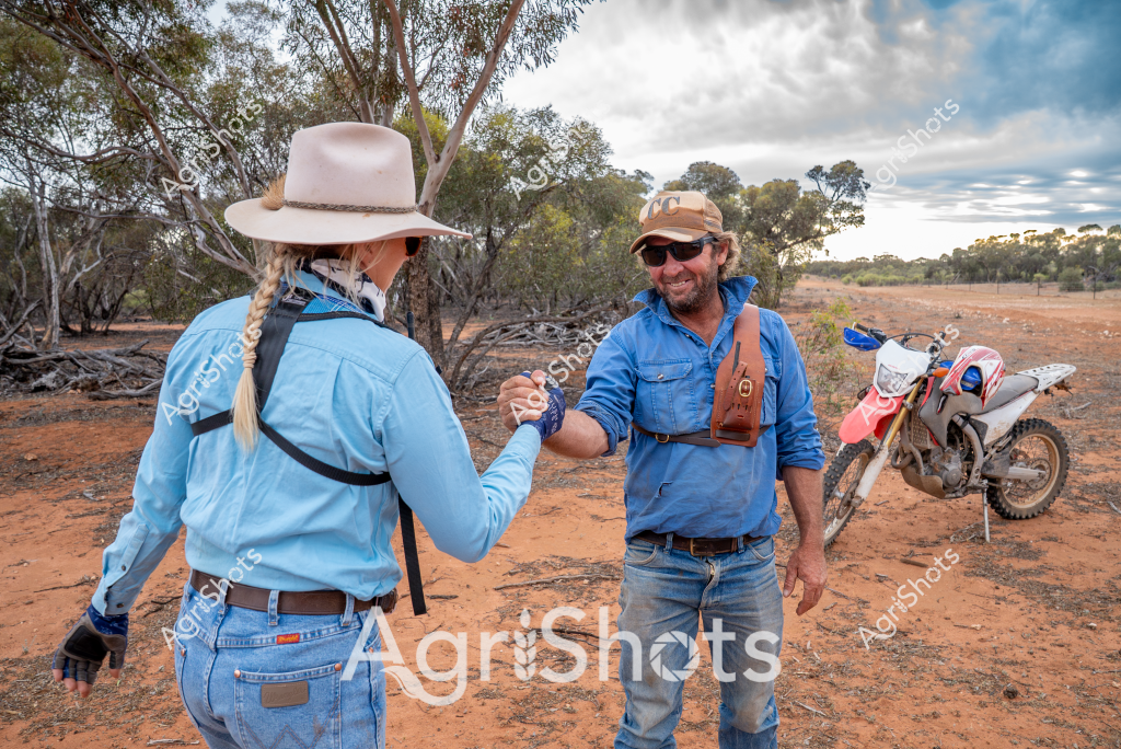 Two people stand in an arid Australian landscape, exchanging a friendly handshake or greeting. A woman wearing a wide-brimmed white hat and light blue work shirt faces a man in similar blue denim clothing, a tan cap, and sunglasses. Behind them, a dirt bike is parked near some scattered eucalyptus trees, suggesting they are likely rural workers or farmers conducting business in the rugged outback environment. The reddish-brown soil and sparse vegetation indicate this is likely a drought-affected region in inland Australia, with a cloudy sky adding to the atmospheric conditions. Their matching blue work shirts and relaxed body language suggest a professional relationship, possibly related to agricultural work or land management in a challenging rural setting.