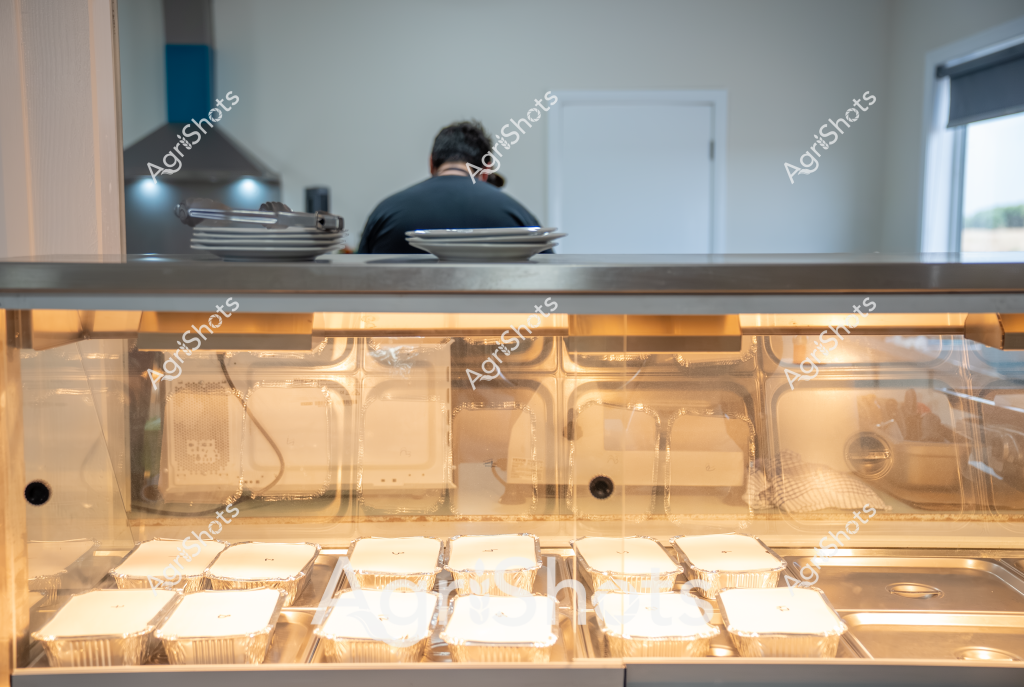 A commercial kitchen workspace is revealed through a service counter, with a person in a dark shirt working at the back preparing food. In the foreground, a professional food display case contains multiple aluminum takeaway containers neatly arranged in rows, illuminated by warm lighting that highlights the pristine food preparation area. The stainless steel countertop and clear display case suggest a modern, hygienic food service environment, likely a restaurant, café, or catering kitchen. Overhead lighting and a window provide ambient illumination, creating a clean and organized workspace. The scene captures the behind-the-scenes precision of food preparation, with carefully arranged meal containers ready for service.