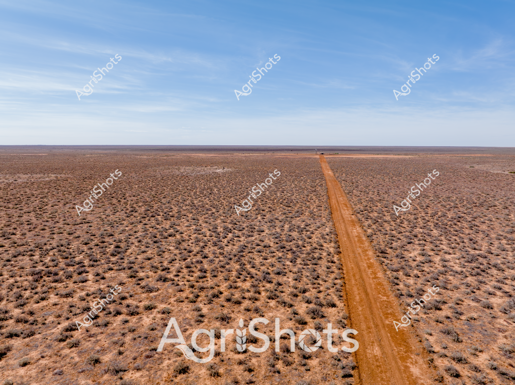 A stark aerial view captures the expansive, arid landscape of the Australian interior, where a long, rust-colored dirt track cuts a perfectly straight line through a sparse, rocky terrain. The landscape is dominated by reddish-brown earth scattered with low, weathered shrubs, creating a textured surface that stretches to the horizon. Wispy clouds drift across a pale blue sky, emphasizing the immense emptiness and isolation of this remote region. The dirt road provides the only visible sign of human intervention, stretching like a precise line drawn across the rugged, drought-affected landscape. The image powerfully illustrates the challenging environmental conditions of Australia's outback, where vast, seemingly endless spaces are punctuated only by the most minimal signs of human presence or infrastructure.

The scene evokes the resilience required to traverse and survive in such a harsh, unforgiving environment, where the landscape itself becomes a testament to the extreme conditions of Australia's interior regions.