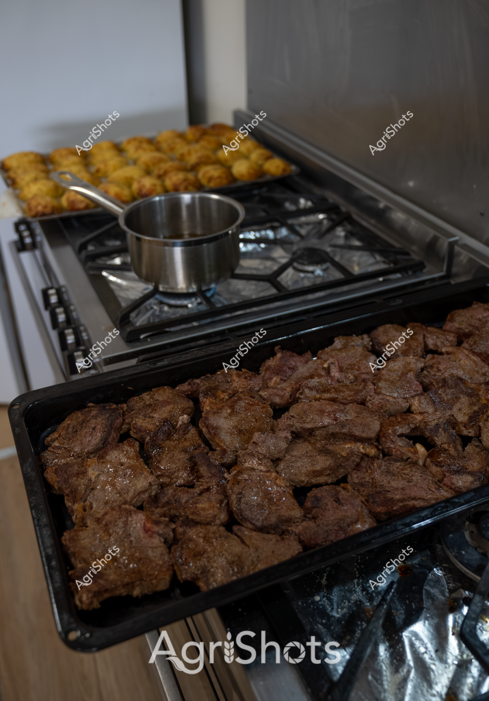 The image depicts a home kitchen stovetop scene focused on meat preparation, with two baking trays showing different stages of cooking. In the foreground, a dark baking tray is filled with browned meat pieces - likely beef chunks or steak cuts that have been seared to a rich, caramelized brown color. On the upper tray, golden-brown roasted or baked items (potentially potato or vegetable side dishes) complement the meat. A stainless steel saucepan sits on the gas stovetop, suggesting ongoing cooking or sauce preparation. The close-up perspective highlights the textural details of the cooked meats, with their crisp exterior and dense, substantial appearance indicating careful culinary preparation. The kitchen setting appears domestic and functional, with a professional-quality cooking approach evident in the precise arrangement and careful browning of the ingredients.