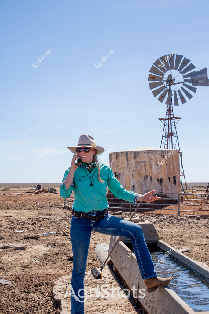 A woman stockman stands confidently beside a water trough in an arid rural landscape, capturing the essence of Australian outback farming. Wearing a turquoise work shirt, blue jeans, a wide-brimmed hat, and sunglasses, she speaks on a mobile phone while leaning against a concrete water channel. Behind her, a classic windmill rises against a clear blue sky, its metal blades silhouetted against the horizon. A large concrete water storage tank stands nearby, typical of remote agricultural infrastructure designed to support livestock in harsh, dry conditions. The ground is dusty and barren, with rocky terrain and minimal vegetation, highlighting the challenging environment of this rural property. Her relaxed posture and professional attire suggest she is managing or monitoring agricultural operations in this remote and unforgiving landscape.
