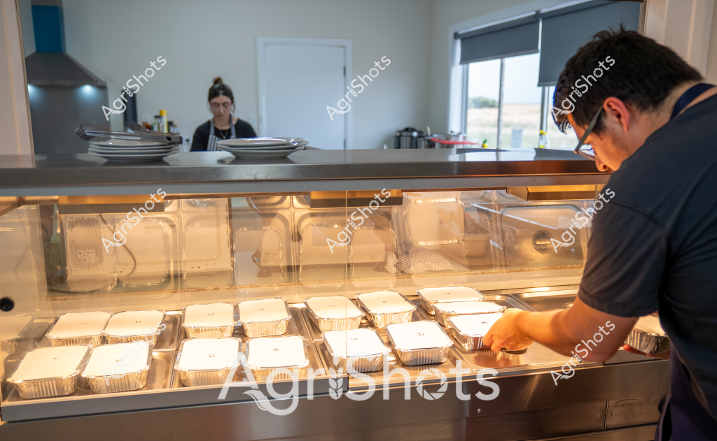 A man in a dark uniform carefully arranges pre-prepared meals in aluminum takeaway containers inside a commercial kitchen display cabinet. The stainless steel food preparation area features a clean, professional workspace with a woman working in the background near kitchen equipment. Neatly organized meal containers are precisely positioned under warm display lighting, suggesting a commercial catering or food service operation. The kitchen appears modern and well-organized, with a sleek counter separating the preparation area from the service zone. Soft natural light filters through a window, adding a bright, clean atmosphere to the food preparation environment.