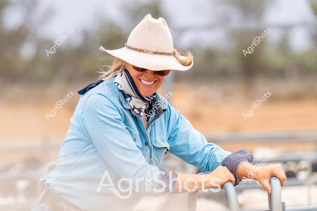 A woman stockman wearing a light blue long-sleeved shirt and a cream-colored cowboy hat leans on a metal gate, smiling broadly in a rural Australian landscape. Her neck is adorned with a patterned scarf, and she's wearing work gloves, suggesting she's in the midst of daily farm activities. The background reveals a soft, blurred rural setting with trees and dry grasslands, typical of an Australian pastoral scene. Her relaxed posture and wide smile convey a sense of contentment and connection to the land, embodying the resilient spirit of rural Australian agricultural workers. The soft lighting and her casual yet purposeful stance capture a moment of quiet satisfaction in the working day of a professional in the agricultural sector.