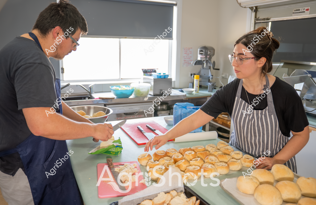 Two people work together in a professional kitchen, preparing a batch of freshly baked bread rolls. A woman wearing a striped apron and glasses stands beside a man, both focused on preparing the golden-brown bread rolls arranged neatly on trays. They are working at a clean, well-lit kitchen workspace with stainless steel equipment and a window in the background. The bread rolls appear uniform in size and color, suggesting careful preparation and baking skills. Their coordinated movements and professional attire indicate they are likely culinary workers or bakers collaborating on food production.