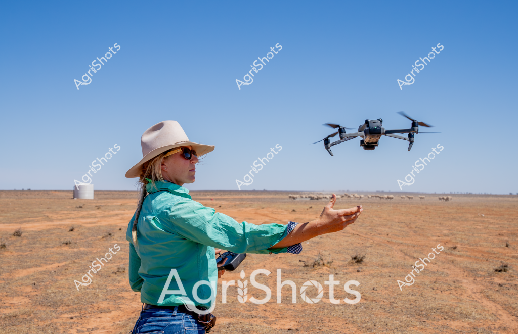 A woman in a turquoise shirt and wide-brimmed sun hat operates a drone in an arid, expansive Australian agricultural landscape. Standing amid a dry, reddish-brown paddock with scattered sparse vegetation, she launches a quadcopter drone with an outstretched arm, controlling its flight with precision. A white water tank stands in the distant background, highlighting the remote rural setting characteristic of Australia's inland regions. The bright blue sky and ochre-colored earth create a stark contrast, emphasizing the challenging environmental conditions typical of agricultural zones during dry periods. The scene captures the intersection of modern agricultural technology and traditional farming practices, with the drone likely being used for land surveying, crop monitoring, or livestock tracking in this remote agricultural environment.